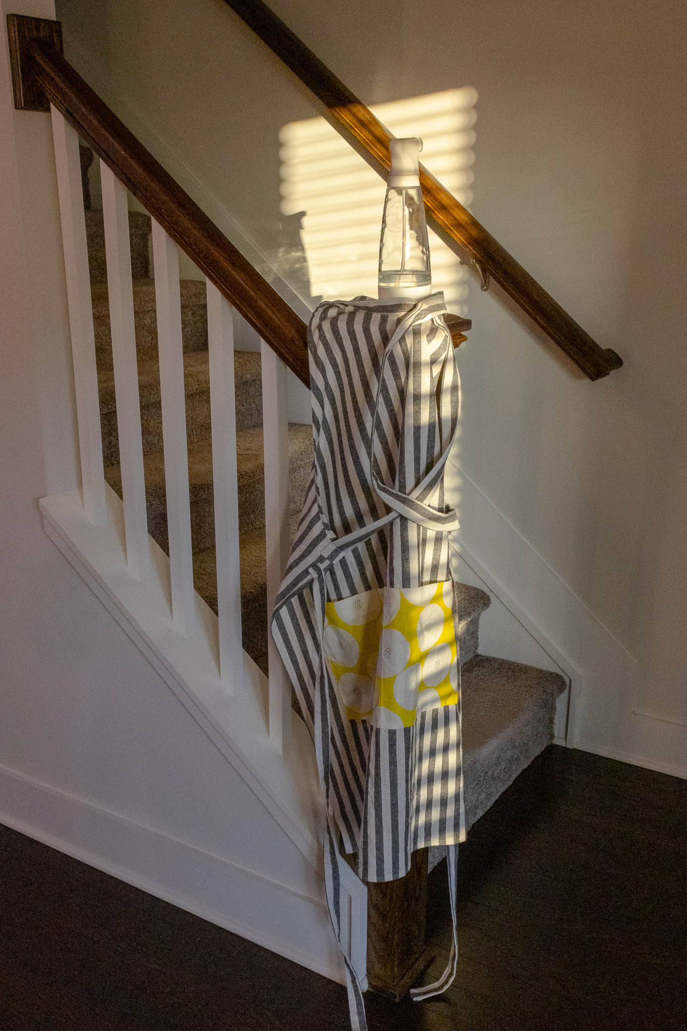 A vertically oriented indoor photograph captures the bottom of a carpeted staircase. Draped over the prominent wooden newel post is an apron with grey and white vertical stripes. The apron features a large, square pocket in a contrasting bright yellow fabric, patterned with large white circles or stylized flowers. Balanced on the flat top of the newel post, nestled within the folds of the apron, sits a clear spray bottle with a white spray mechanism, appearing to contain a clear liquid. Sunlight streams in from the right, casting distinct horizontal stripes of light and shadow across the off-white wall behind the stairs, the dark wood handrail ascending diagonally, and parts of the apron and spray bottle. This suggests the light is coming through window blinds. The staircase itself has white balusters (spindles) and light grey or beige carpeting on the treads. The flooring at the bottom of the stairs is a dark color. The overall composition focuses on the apron and bottle on the newel post, bathed in the striped sunlight.